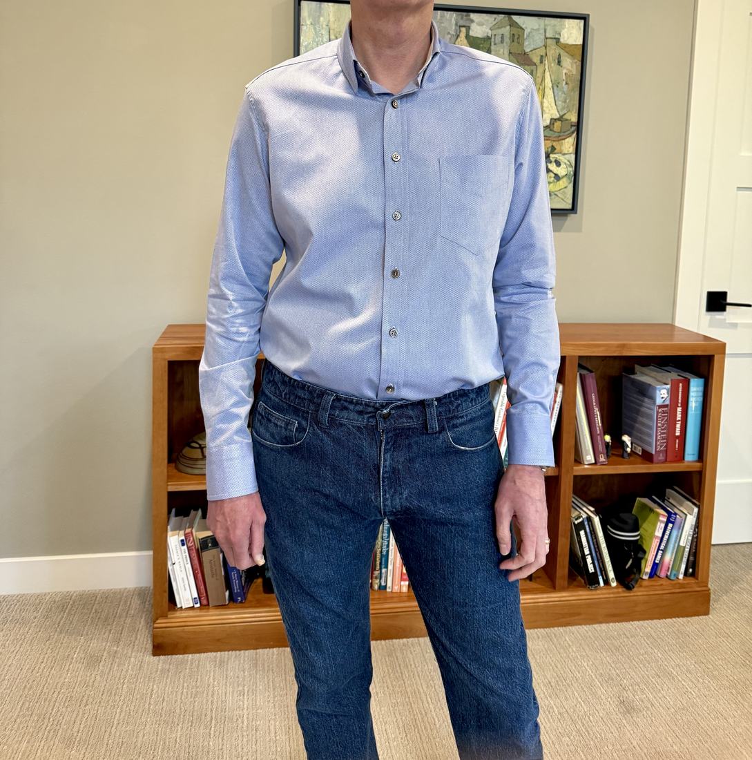 The finished blue dress shirt worn with jeans in front of a low bookcase.