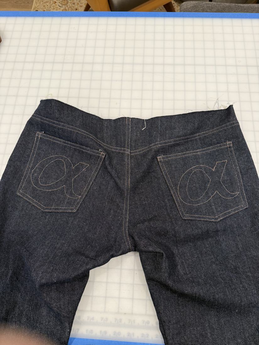 A pair of raw denim jeans laid flat, showing both embroidered back pockets.
