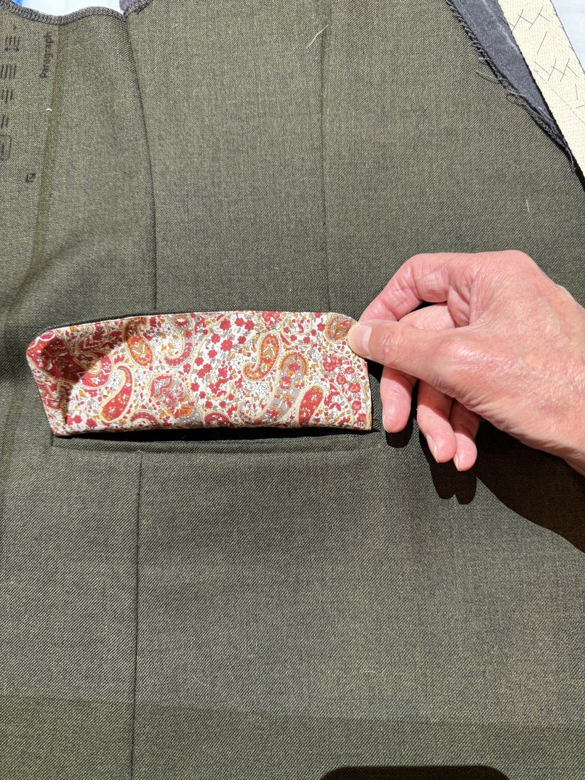 A welt pocket in olive wool faced in red paisley, held open by hand.