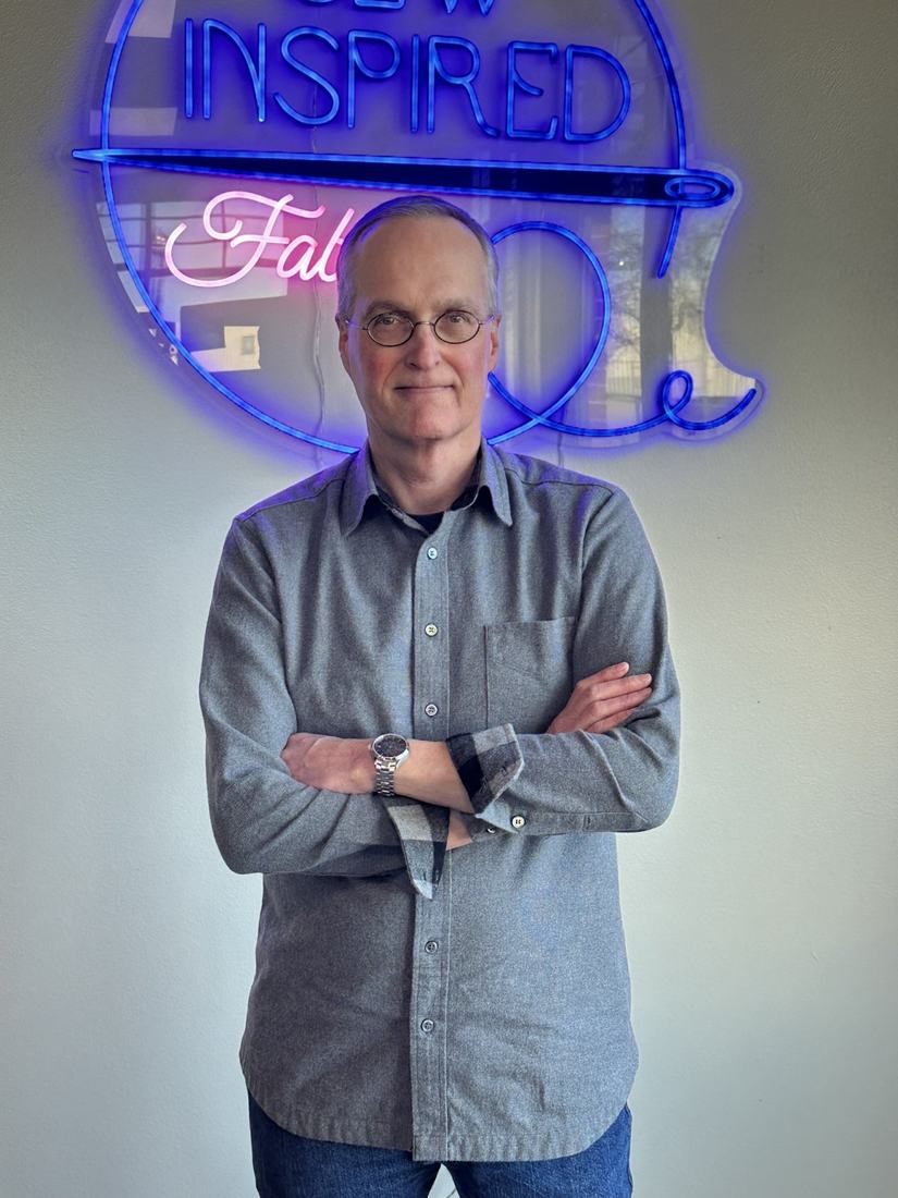 Standing with arms crossed in a grey flannel shirt in front of a neon Sew Inspired Fabrics sign.